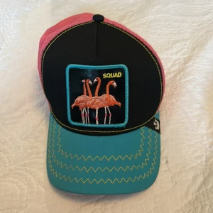 Färgglad keps med flamingo-brodyr från The Farm  - Keps med svart front, rosa mesh baktill och turkos skärm. Broderad patch med flamingos och texten 'SQUAD' framtill. Dekorativa gula sömmar på skärmen. Justerbar baktill.