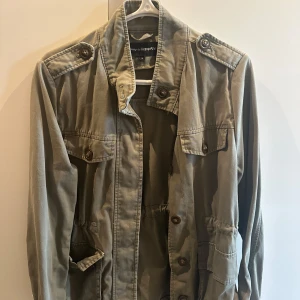 Olivgrön utilityjacka med knappar - Säljer en olivgrön utilityjacka från KappAhl med flera fickor och dekorativa axelpartier. Jackan har knappar framtill och på fickorna samt lång ärm. Perfekt för lager-på-lager under svalare dagar.