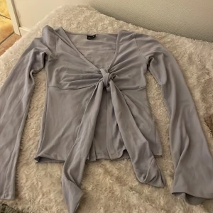 Ljusblå långärmad topp med knyt - Säljer en ljusblå långärmad topp med djup v-ringning och knyt framtill. Toppen har en figurnära passform och är i ett mjukt, stretchigt material. Den är använd lite så den är lite nopprig men annars är den i toppenskick. 