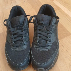 Svarta Nike sneakers - Säljer ett par helsvarta Nike sneakers med klassisk design och snörning. Skorna har rund tå och platt sula, tillverkade i en mix av syntet och textil. Perfekta för dig som gillar stilrena och bekväma skor till vardags.