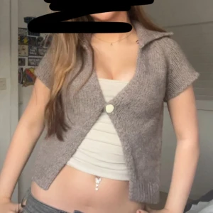Grå stickad kofta med kort ärm och krage - Säljer en grå stickad kofta med korta ärmar och stor krage. Koftan är croppad och stängs med en knapp framtill. Perfekt för lager-på-lager och ger en chill vibe till outfiten.