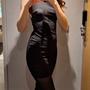 Svart tight klänning med korsade band och dragkedja - Säljer en svart tight klänning med korsade band i ryggen och guldfärgad dragkedja. Klänningen är ärmlös och har en kroppsnära passform som framhäver figuren. Perfekt för dig som vill ha en stilren och modern look.