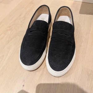 Marinblå mockaloafers med vit sula stl 42 - Snygga marinblå loafers i mocka med vit platt sula från Goosecraft. Skorna har en klassisk design med rund tå och diskreta detaljer på ovandelen. Perfekta för en stilren och avslappnad look.