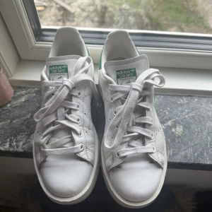 Adidas Stan Smith vita sneakers med gröna detaljer - Klassiska vita Adidas Stan Smith sneakers med gröna detaljer på hälen och tungan. Skorna har snörning, perforerade ränder på sidorna och platt sula. Perfekta för en stilren och sportig look.