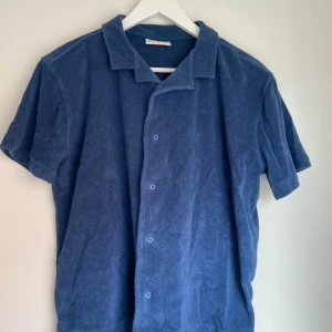 Blå frottéskjorta med kort ärm - Säljer en blå skjorta i mjuk frotté med korta ärmar och klassisk krage. Skjortan har knappar framtill och en avslappnad passform, perfekt för varma dagar. Enkel och stilren design som passar till mycket.