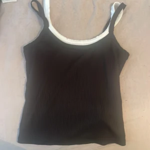 Svart ribbad linnetopp med vita band - Snygg svart ribbad linnetopp med kontrasterande vita axelband och kant. Perfekt för sommardagar och enkel att matcha med jeans eller kjol. Toppens ribbade material ger en trendig känsla.