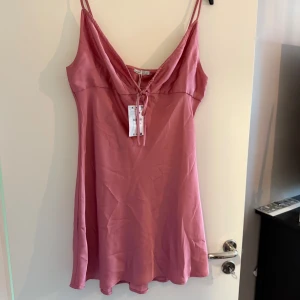 Rosa satin klänning med smala axelband - Säljer en söt rosa klänning i satin med smala axelband och v-ringning. Klänningen har en feminin passform och knyt framtill vid bysten. Perfekt för sommarens alla tillfällen!