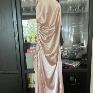 Ljusrosa långklänning med öppen rygg  - Säljer en elegant ljusrosa långklänning i satin med enaxlad design och smala axelband. Klänningen har snygg drapering på sidan och en mjuk glansig yta som ger en lyxig känsla. Perfekt för speciella tillfällen.