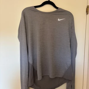 Grå långärmad träningströja från Nike - Säljer en grå långärmad träningströja från Nike med rund hals och vit logga på bröstet. Perfekt för löpning eller gym. Materialet är lätt och andas bra.