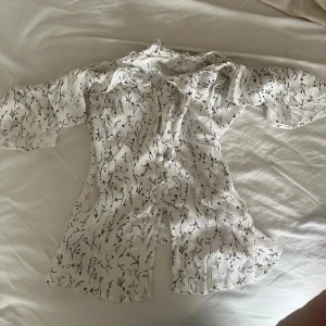 Vit blus med svart mönster och volang - Säljer en vit blus med svart grenliknande mönster och stora volangdetaljer vid axlarna. Blusen har knappar framtill och är tillverkad i ett lätt, luftigt material. Perfekt för dig som gillar unika detaljer och en feminin stil. Sitter som S