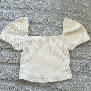 Beige ribbad topp med puffärmar - Snygg beige ribbad topp med korta puffärmar och fyrkantig halsringning. Perfekt för en trendig och stilren look. Mjuk och stretchig i materialet.