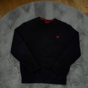 Mörkblå sweatshirt från Hugo - Säljer en mörkblå sweatshirt från Hugo med rund halsringning och diskret röd logotyp på bröstet. Tröjan har långa ärmar och är tillverkad i mjuk bomull. Den är i storlek M men passar mer som S. Perfekt för en stilren och enkel look.