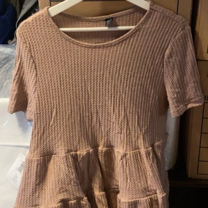 Beige kortärmad blus med struktur - Säljer en beige bjud med kort ärm och rund halsringning. Blusen har en strukturerad, quiltad yta och volang nedtill för en avslappnad look. Perfekt för dig som gillar bekväma och stilrena plagg.