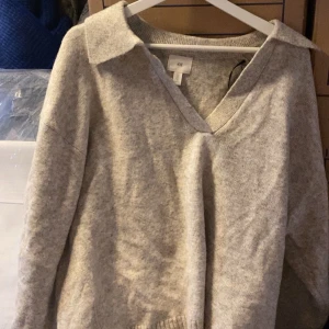 Ljusgrå stickad tröja från H&M - Mysig ljusgrå stickad tröja från H&M med v-ringning och krage. Tröjan har lång ärm och en avslappnad passform, perfekt för lager på lager under kyliga dagar.