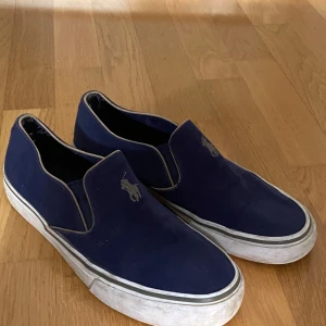 Blå slip-on sneakers från Polo Ralph Lauren - Mörkblå slip-on sneakers med vit sula och Polo Ralph Lauren-logga broderad på ovansidan. Skorna har en enkel och stilren design utan snörning, perfekt för en avslappnad look. Perfekta till sommaren!