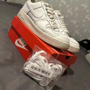 Vita Nike Air Force 1 sneakers - Klassiska vita Nike Air Force 1 sneakers med låg profil och rund tå. Skorna har snörning och är tillverkade i skinn med en platt sula. Slitna vid hälen men går fortfarande bra att använda. Levereras med extra vita skosnören. Perfekta för dig som gillar en clean och tidlös stil.