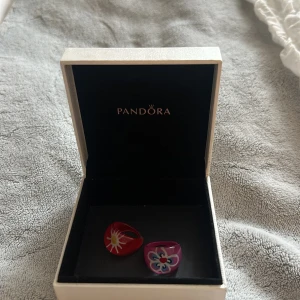 Två färgglada ringar från Pandora - Säljer två unika ringar från Pandora i röd och lila färg. Den röda ringen har ett solmönster i vitt och gult, medan den lila ringen har ett blommönster i blått, vitt och rosa. Perfekta för att ge din stil en färgklick!