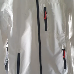 Vit vindjacka från Helly Hansen - Säljer en vit vindjacka från Helly Hansen med svarta och röda detaljer på dragkedjorna. Jackan har lång ärm, flera fickor med dragkedja och meshfoder på insidan. Perfekt för blåsiga dagar och outdoor-aktiviteter.