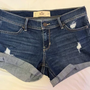 Mörkblå jeansshorts från Hollister - Snygga mörkblå jeansshorts från Hollister med upprullade benslut och slitna detaljer framtill. Klassisk femficksmodell med knapp och dragkedja. Perfekta för sommaren och har en bekväm midwaist-passform.