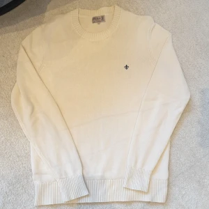 Beige stickad tröja från Morris - Säljer en stilren vit stickad tröja från Morris med rund halsringning och diskret broderad logga på bröstet. Tröjan har långa ärmar och breda ribbade muddar vid ärmslut och nederkant. Perfekt för en klassisk look. För ändast 600kr.