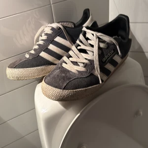 Adidas Gazelle sneakers i svart mocka - Säljer ett par klassiska Adidas Gazelle sneakers i svart mocka med vita ränder och vit sula. Skorna har snörning och en platt sula, perfekt för dig som gillar retrostil. Ikonisk Adidas-logga på sidan och tungan.