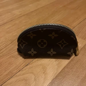 Brun monogram plånbok - Säljer en liten brun plånbok med klassiskt monogrammönster och guldfärgad dragkedja. Perfekt för kort eller småmynt. Smidig att ha med sig i väskan eller fickan.