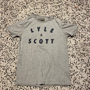 Grå t-shirt från Lyle & Scott Junior - Snygg grå t-shirt från Lyle & Scott Junior med stor mörkblå logga och text på bröstet. Klassisk rund halsringning och korta ärmar. Perfekt för en avslappnad stil.