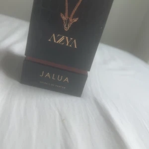 Azya Jalua Extrait de Parfum - Lyxig parfymflaska från Azya med doften Jalua. Flaskan är genomskinlig med guldigt lock och innehåller en gyllengul vätska. Förpackningen är svart med gulddetaljer och har en elegant design med ett djurmotiv på framsidan.