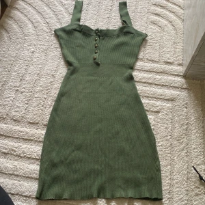 Grön ribbad klänning med knappar - Säljer en grön ribbad klänning med breda axelband och dekorativa knappar framtill. Klänningen har en figurnära passform och är perfekt för sommardagar. Den är stretchig och bekväm med en fin struktur.