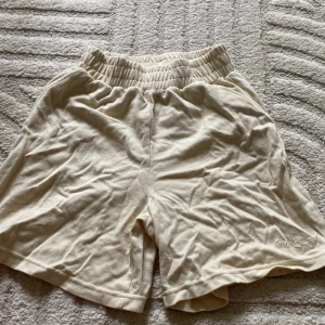 Beige shorts från Oh Polly - Beiga shorts från Oh Polly med resår i midjan och broderad logga 'Polly Sport' på benet. Tillverkade i mjuk bomull, perfekta för en avslappnad stil. Klassisk passform och enkel design.