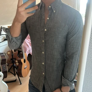 Grå skjorta från Ralph Lauren - Säljer en stilren grå skjorta från Ralph Lauren med klassisk button-down krage och broderad logga på bröstet. Skjortan har långa ärmar och vita knappar framtill. Perfekt till jeans eller chinos.