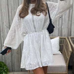 Student klänning: Vit kortklänning med broderade detaljer - Superfin vit klänning med broderade detaljer och lös passform. Klänningen har långa, vida ärmar och en markerad midja med resår. Dekorativ spets längs mitten framtill. Perfekt för sommarens alla tillfällen! Passar allt från xs-m då den är justeringsbar