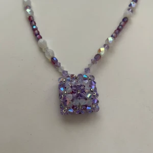 Lila och vit glaspärlhalsband med kubhänge - Unikt halsband med små glaspärlor i lila, vitt och iriserande nyanser. Halsbandet har ett handgjort kubformat hänge av lila och genomskinliga pärlor. Låset är i silverfärgad metall med blommönster. Perfekt för dig som gillar färgglada accessoarer!