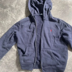 Mörkblå hoodie från Polo Ralph Lauren - Säljer en mörkblå hoodie från Polo Ralph Lauren. Klassisk design med röd broderad logga på bröstet och två fickor framtill. Tillverkad i mjuk bomull och polyesterblandning,