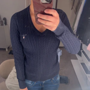 Mörkblå kabelstickad tröja från GANT - Säljer en stilren mörkblå kabelstickad tröja från GANT med v-ringning. Tröjan har långa ärmar och ett broderat märke på bröstet. 