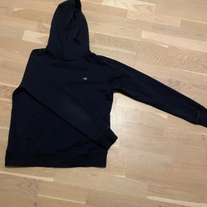 Mörkblå  hoodie från GANT - Säljer en stilren mörkblå hoodie från GANT med en liten broderad logotyp på bröstet. Perfekt för en avslappnad look. Den har en klassisk huva och långa ärmar.