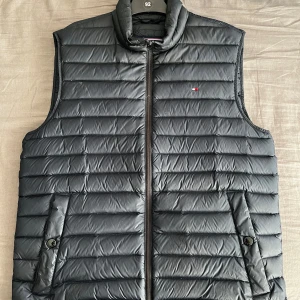 Mörkblå väst från Tommy Hilfiger - Säljer en stilren mörkblå dunväst från Tommy Hilfiger. Västen har en dragkedja framtill och två fickor med knappar. Perfekt för lager-på-lager under kyligare dagar. Klassisk design med diskret logga på bröstet. Ställ gärna fler frågor! Endast Swish betalning!!!
