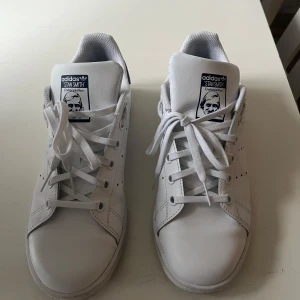 Vita Adidas Stan Smith sneakers - Klassiska vita Adidas Stan Smith sneakers med mörkblå detalj på hälen. Skorna har en stilren design med perforerade ränder och snörning framtill. Perfekta för en avslappnad och trendig look. Testade ute en gång! Nypris ca 1200kr