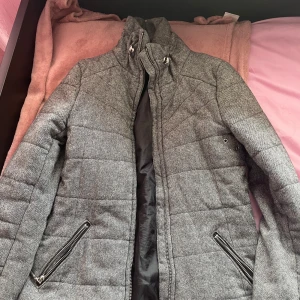 Grå quiltad jacka från H&M - Säljer en stilren grå quiltad jacka från H&M. Jackan har en hög krage och dragkedja framtill samt två fickor med dragkedjor. Perfekt för kyligare dagar.