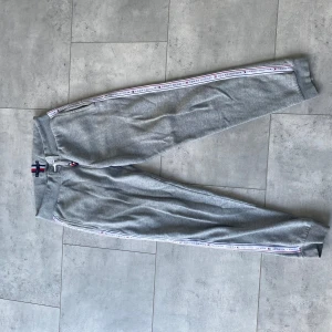 Grå mjukisbyxor från Tommy Hilfiger - Säljer ett par grå mjukisbyxor från Tommy Hilfiger med ribbade muddar och snörning i midjan. Byxorna har en dekorativ rand med logotyp längs sidorna. Perfekta för en avslappnad stil.