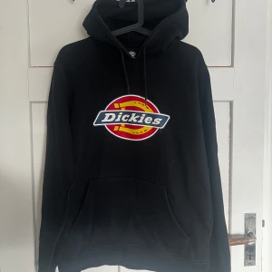 Svart hoodie från Dickies - Säljer en svart hoodie från Dickies med deras ikoniska logga i rött, gult och blått på bröstet. Tröjan har en klassisk känguruficka och justerbar huva med dragsko. Perfekt för en avslappnad stil.