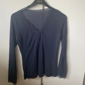 Mörkblå långärmad topp från intimissmi  - Snygg mörkblå topp med långa ärmar och v-ringning. Toppen har fina spetsdetaljer vid halsringningen och ärmsluten, vilket ger en elegant touch. Perfekt för att klä upp en vardagsoutfit eller för en kväll ute.
