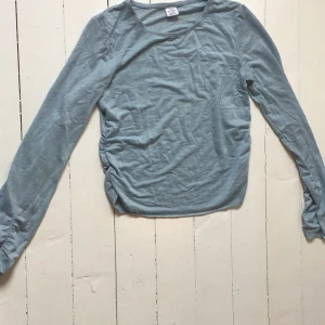 Blå glittrig tröja från Lindex - Säljer en blå glittrig tröja från Lindex med långa ärmar och rynkade detaljer på sidorna. Perfekt för att lägga till lite glitter i vardagen. Tröjan har en rund halsringning och är tillverkad i ett lätt material.