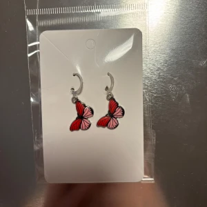 Fjärilsörhängen i rött och rosa - Charmiga örhängen med fjärilsdesign i rött och rosa. Perfekta för att lägga till en färgglad touch till din outfit. De hänger från en silverfärgad krok och är lätta att bära. (Billigt material) 