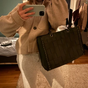 Grön axelväska med flätad design - Snygg grön axelväska med en unik flätad design och nitar längs kanterna. Väskan har en praktisk dragkedja på baksidan och robusta handtag. Perfekt för att bära dina dagliga nödvändigheter med stil.