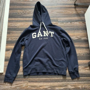 Mörkblå hoodie från GANT - Säljer en snygg mörkblå hoodie från GANT med vit text på bröstet. Tröjan har en klassisk design med huva och dragsnören. Perfekt för en avslappnad stil.