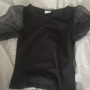 Svart topp med puffärmar från Vila - Snygg svart topp från Vila med puffiga, transparenta ärmar. Perfekt för en stilren look med en twist. Toppen har en rund halsringning och är tillverkad i ett mjukt material. Köpt ny för 400