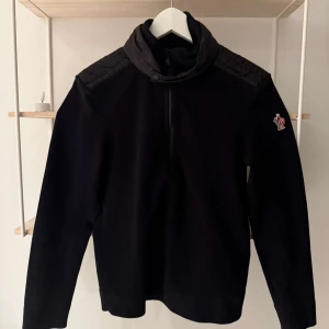 Svart tröja med dragkedja från Moncler Storlek S - Säljer en stilren svart tröja från Moncler med dragkedja och hög krage. Tröjan har ett broderat märke på ärmen och quiltade detaljer på axlarna. Perfekt för kyliga dagar.
