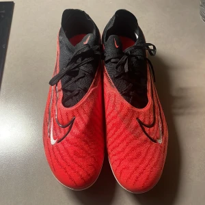 Röda Nike phantom fotbollsskor - Ett par Nike Phantom GX Elite SG-Pro AC. Snygga röda fotbollsskor från Nike med svart snörning och vit sula. Skorna har en modern design med svarta och vita detaljer samt en mönstrad yta. Perfekta för fotbollsplanen!