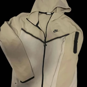 Beige hoodie från Nike 250kr - Säljer en beige hoodie från Nike med dragkedja och huva. Jackan har en svart Nike-logga på bröstet och en praktisk ficka med dragkedja på ärmen. Perfekt för en avslappnad stil.